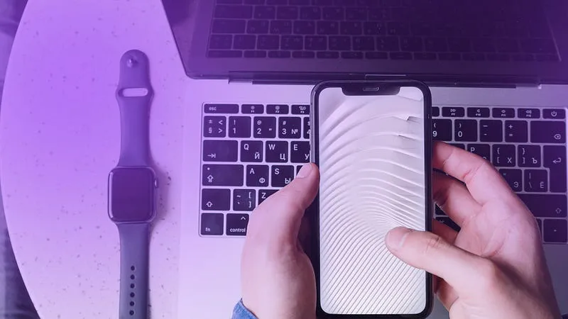 Hands holding a smartphone above a laptop keyboard next to a smartwatch, illustrating mobile haptics