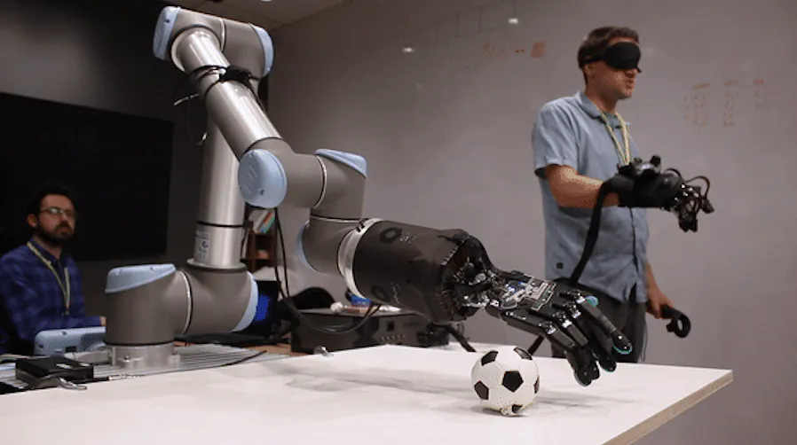 Operator wearing haptic gloves remotely controlling a robotic arm and hand reaching for a soccer ball