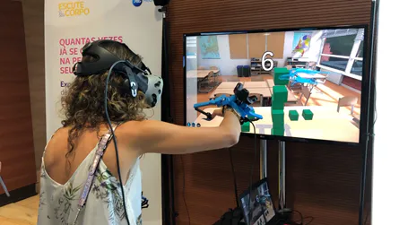 Visitor in a VR headset using hand controllers at a booth demo, the simulation shown on a screen behind